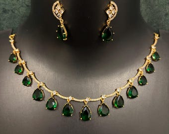Puede incluir: Un conjunto de collar y pendientes en tono dorado con piedras de color verde oscuro en forma de lágrima. El collar tiene una cadena delicada con pequeños cristales transparentes y varios colgantes verdes. Los pendientes a juego tienen un diseño similar.
