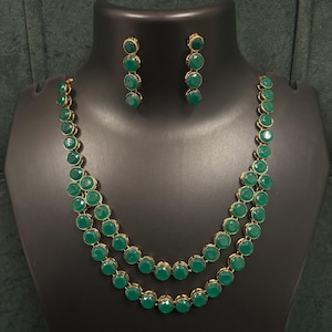 Pode incluir: Um conjunto de colar e brincos a condizer. A joia apresenta grandes pedras redondas verde esmeralda engastadas em metal dourado. O colar tem duas voltas. O conjunto é exibido num manequim preto.