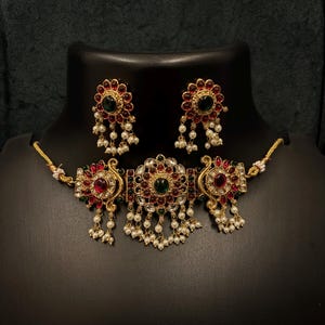 Puede incluir: Un conjunto de collar y pendientes en tono dorado. El collar presenta un diseño central con piedras preciosas rojas y verdes, rodeado de pequeñas perlas blancas. Los pendientes reflejan el diseño, con un patrón floral y perlas colgantes. La joyería se exhibe en un maniquí negro.