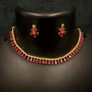 May include: A gold-toned necklace and matching earrings set. The necklace features a row of small, red teardrop-shaped stones and a delicate chain. The earrings are flower-shaped with red petals. The jewelry is displayed on a black mannequin.
