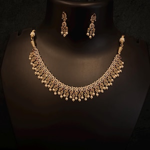 May include: A gold-coloured necklace and matching earrings set displayed on a black mannequin. The necklace features ornate, circular designs with red and clear stones, and dangling white pearls. The earrings mirror the necklace design.