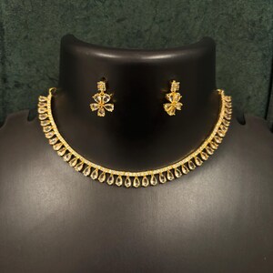 May include: A gold-coloured necklace and matching earrings set. The necklace has a delicate chain with teardrop-shaped pendants. The earrings and necklace have floral designs with clear, sparkling stones. The jewellery is displayed on a black mannequin.