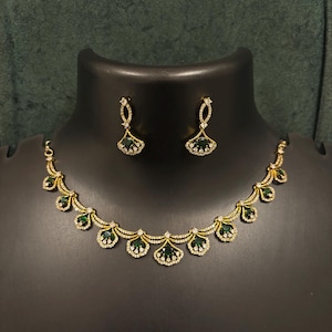 Puede incluir: Un conjunto de collar y pendientes dorados. El collar presenta una serie de piedras verdes esmeralda en forma de abanico, cada una acentuada con pequeños cristales transparentes. Los pendientes a juego tienen un diseño similar.