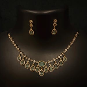 Op de afbeelding: Een goudkleurige ketting- en oorbellenset. De ketting heeft een centrale ovale groene edelsteen, omringd door kleinere heldere stenen. Druppelvormige groene edelstenen hangen eronder. De oorbellen hebben een vergelijkbaar ontwerp.