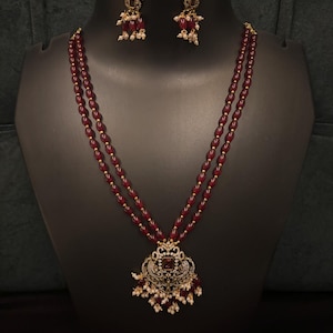 Peut inclure: Ensemble de bijoux composé d'un collier et de boucles d'oreilles assorties. Le collier est orné de perles bordeaux et d'un pendentif doré avec une pierre rouge et des perles blanches. Les boucles d'oreilles ont un design similaire.