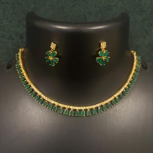 May include: A gold-toned necklace and earring set. The necklace features a row of green teardrop-shaped stones and a matching pair of earrings with green floral designs. The jewelry is displayed on a black mannequin.