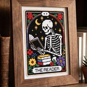 K&ouml;nnte beinhalten: Gerahmtes Kreuzstichmuster mit einem Skelett, das ein Buch liest, vor schwarzem Hintergrund mit floralen Akzenten. Der Text "THE READER" steht unten. Der Rahmen ist aus Holz. Die Gr&ouml;&szlig;e betr&auml;gt 105 x 147 Stiche.