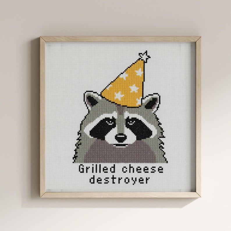 Op de afbeelding: Kruissteekkunst van een wasbeer met een gele feesthoed met witte sterren. De wasbeer heeft een zwart masker en is ingelijst in een houten vierkant. De tekst "Grilled cheese destroyer" staat eronder.