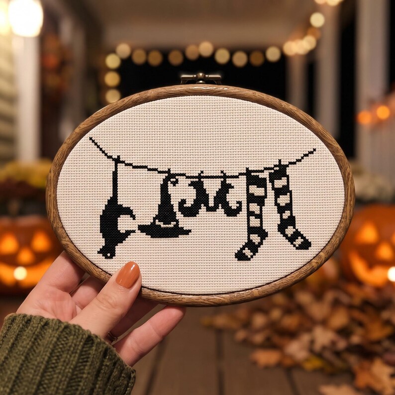 May include: An oval wooden embroidery hoop containing white fabric with a black cross-stitch design. The pattern depicts a clothesline with a cat silhouette, a witch hat, a pair of boots, and striped stockings hanging from a thin black string.
