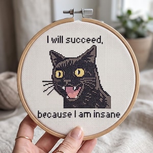 May include: A circular wooden embroidery hoop containing white fabric with a cross-stitch design of a black cat with yellow eyes and an open mouth showing teeth. The text above reads I will succeed, and below reads because I am insane.