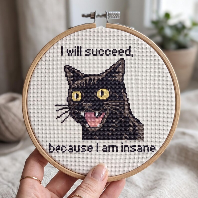 May include: A circular wooden embroidery hoop containing white fabric with a cross-stitch design of a black cat with yellow eyes and an open mouth showing teeth. The text above reads I will succeed, and below reads because I am insane.
