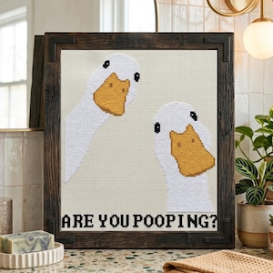 May include: Cross-stitch artwork featuring two white geese with orange beaks and black eyes. The phrase "ARE YOU POOPING?" is stitched below the geese in black letters. The artwork is framed in a dark brown, rustic wooden frame.