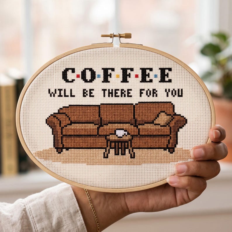 May include: An oval embroidery hoop containing a cross-stitch design on cream fabric. The text reads COFFEE WILL BE THERE FOR YOU in black, with colorful dots between letters. Below, a brown sofa and a small table with a white cup are stitched.