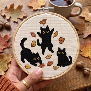 May include: A circular wooden embroidery hoop containing a cross-stitch design on cream fabric. The design features three black cats with large yellow eyes surrounded by scattered autumn leaves in shades of orange, yellow, and brown.