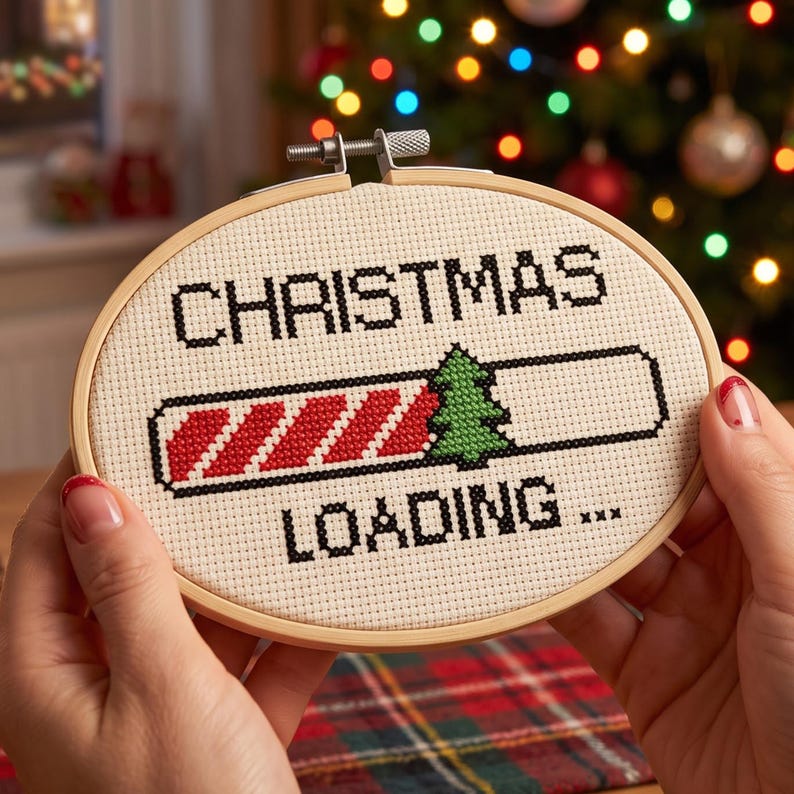 Pu&ograve; includere: Un telaio da ricamo ovale in legno contenente un disegno a punto croce su tessuto color crema. Il testo recita CHRISTMAS LOADING con una barra di caricamento a strisce diagonali rosse e bianche e un'icona di pino verde.