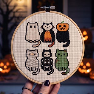 Puede incluir: Un bastidor de bordado circular de madera que contiene un patr&oacute;n de punto de cruz de seis gatos con disfraces de Halloween. Los gatos est&aacute;n representados como un fantasma, vampiro, calabaza, momia, esqueleto y monstruo verde en tela blanca.