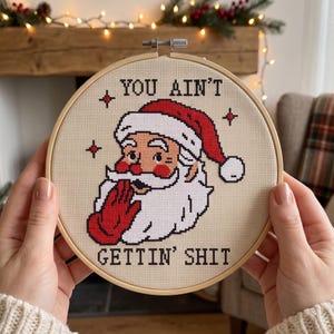 May include: A circular wooden embroidery hoop containing a cross-stitch design of Santa Claus wearing a red hat and glove, with the black text "YOU AIN'T GETTIN' SHIT" stitched above and below the figure on a light beige fabric background.