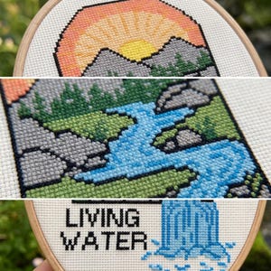 K&ouml;nnte beinhalten: Eine Kreuzstichstickerei in einem Holzrahmen mit einer Landschaft aus gelber Sonne, orangefarbenem Himmel, grauen Bergen, gr&uuml;nen B&auml;umen und einem blauen Fluss. Der untere Teil zeigt einen blauen Wasserfall und den schwarzen Text LIVING WATER.