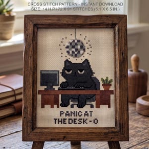Puede incluir: Un patr&oacute;n de punto de cruz con un gato negro sentado en un escritorio marr&oacute;n con un ordenador y una bola de discoteca. El texto dice PANIC AT THE DESK-O. El patr&oacute;n es de 72 x 91 puntos, 14 H.P.I, mide 13 x 16,5 cm y est&aacute; en un marco.