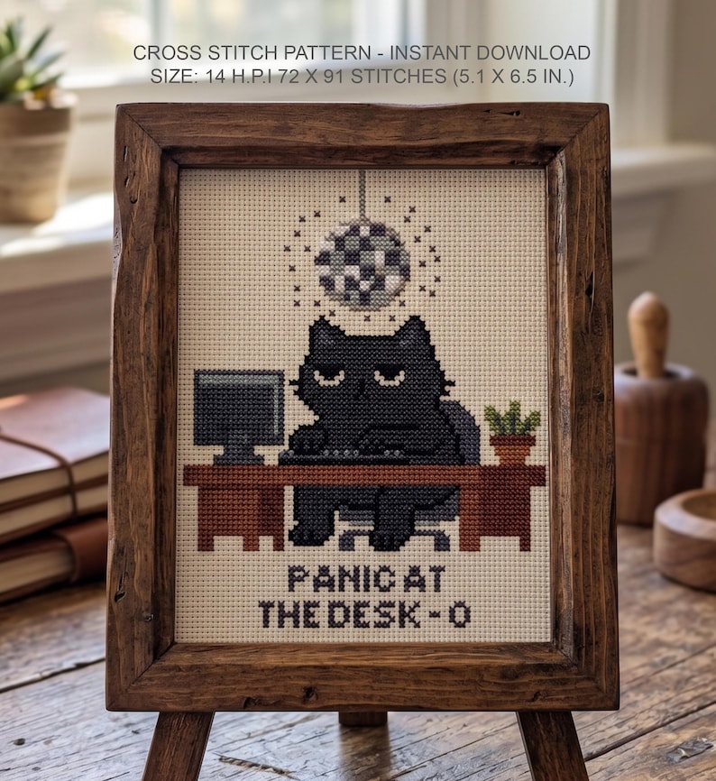 Puede incluir: Un patr&oacute;n de punto de cruz con un gato negro sentado en un escritorio marr&oacute;n con un ordenador y una bola de discoteca. El texto dice PANIC AT THE DESK-O. El patr&oacute;n es de 72 x 91 puntos, 14 H.P.I, mide 13 x 16,5 cm y est&aacute; en un marco.