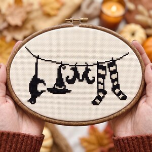 May include: An oval wooden embroidery hoop containing a cross-stitch design on cream fabric. The design features a black thread pattern of a hanging line with a cat silhouette, a witch hat, a bat, and two striped stockings suspended from it.