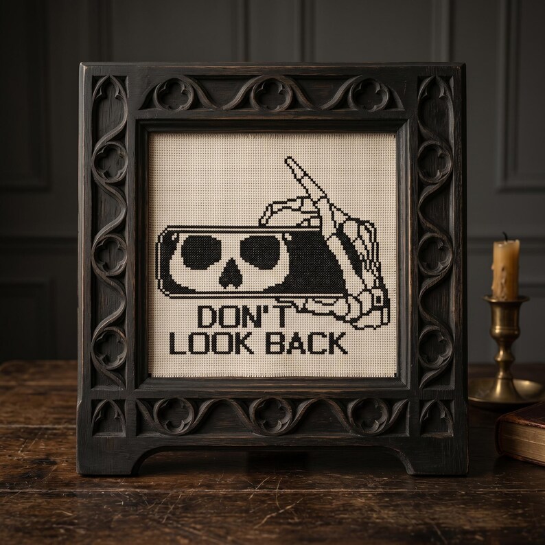 May include: A square cross-stitch embroidery in a dark, ornate wooden frame. The design features a black skeleton hand holding a smartphone displaying a skull icon, with the text DON'T LOOK BACK stitched in black thread on a cream fabric background.