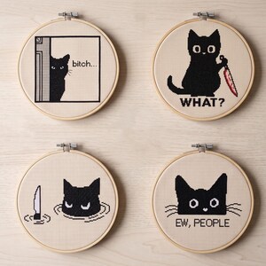 Peut inclure: Quatre tambours &agrave; broder circulaires en bois contenant des points de croix sur tissu beige. Les motifs montrent des chats noirs avec du texte : un qui regarde en disant "bitch...", un tenant un couteau ensanglant&eacute; "WHAT?", un dans l'eau et "EW, PEOPLE".