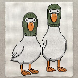 Peut inclure: Une broderie au point de croix sur tissu blanc repr&eacute;sentant deux canards blancs c&ocirc;te &agrave; c&ocirc;te. Chaque canard porte une cagoule vert fonc&eacute; qui ne laisse appara&icirc;tre que leur bec orange et leurs grands yeux ronds blancs &agrave; pupilles noires.