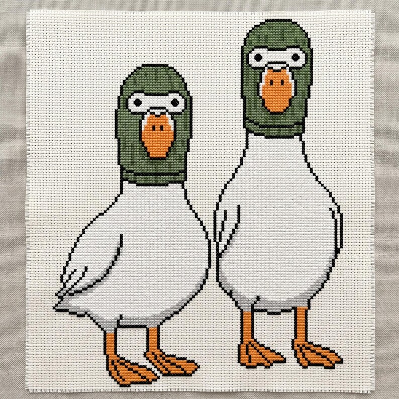 Peut inclure: Une broderie au point de croix sur tissu blanc repr&eacute;sentant deux canards blancs c&ocirc;te &agrave; c&ocirc;te. Chaque canard porte une cagoule vert fonc&eacute; qui ne laisse appara&icirc;tre que leur bec orange et leurs grands yeux ronds blancs &agrave; pupilles noires.