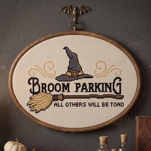 May include: An oval cross-stitch embroidery in a wooden hoop featuring a grey witch hat, a broom, and the text "BROOM PARKING ALL OTHERS WILL BE TOAD". A bronze-coloured metal bat hanger is attached to the top of the wooden embroidery frame.
