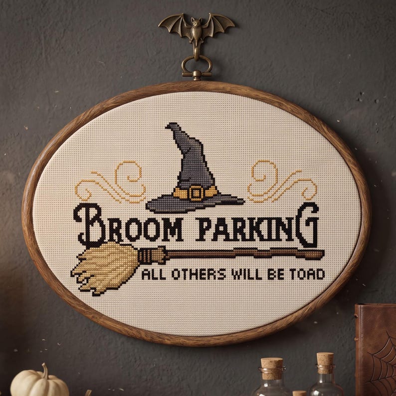 May include: An oval cross-stitch embroidery in a wooden hoop featuring a grey witch hat, a broom, and the text "BROOM PARKING ALL OTHERS WILL BE TOAD". A bronze-coloured metal bat hanger is attached to the top of the wooden embroidery frame.