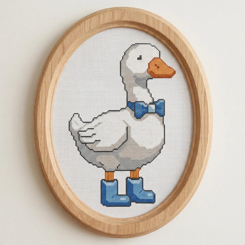 May include: Cross-stitch picture of a white goose wearing a blue bow tie and blue boots, in an oval wooden frame. The goose has an orange beak and is on a white background.