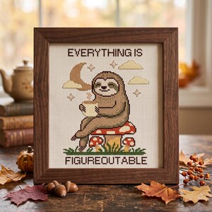 May include: A framed cross-stitch embroidery featuring a brown sloth sitting on a red and white spotted mushroom, holding a mug. The text reads EVERYTHING IS FIGUREOUTABLE. The design includes a crescent moon, clouds, and stars on cream fabric.