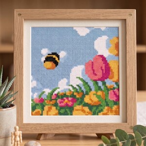 K&ouml;nnte beinhalten: Kreuzstich-Kunstwerk in einem Holzrahmen. Das Bild zeigt eine Biene, die in einem blauen Himmel mit wei&szlig;en Wolken &uuml;ber bunten Blumen fliegt. Die Blumen sind rosa, gelb und orange. Der Rahmen ist hellbraun.
