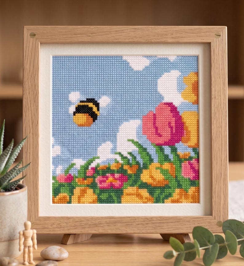 K&ouml;nnte beinhalten: Kreuzstich-Kunstwerk in einem Holzrahmen. Das Bild zeigt eine Biene, die in einem blauen Himmel mit wei&szlig;en Wolken &uuml;ber bunten Blumen fliegt. Die Blumen sind rosa, gelb und orange. Der Rahmen ist hellbraun.