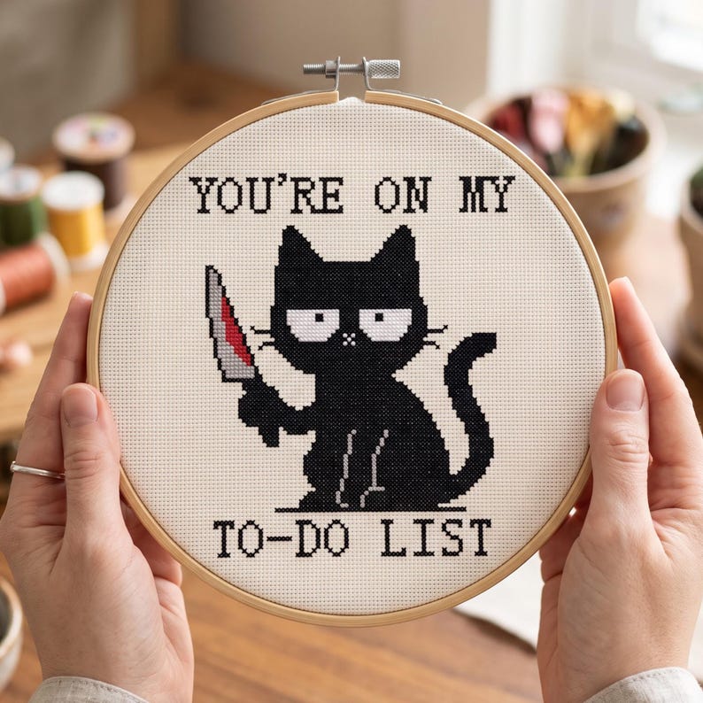 Pu&ograve; includere: Un telaio da ricamo circolare in legno contenente un pezzo di tessuto bianco con un disegno a punto croce di un gatto nero che tiene in mano un coltello. Il testo sopra il gatto recita YOU'RE ON MY e sotto recita TO-DO LIST in nero.