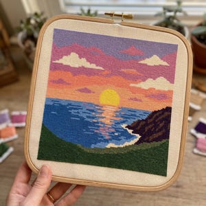 May include: A square cross-stitch embroidery in a wooden hoop featuring a sunset over the ocean. The scene depicts a bright yellow sun, a blue sea with reflections, a dark rocky cliff on the right, and a pink and orange sky with white clouds.