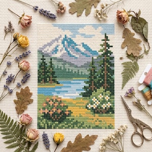 Puede incluir: Un paisaje de punto de cruz con una cordillera, un lago y árboles de hoja perenne. La escena está representada en tonos de azul, verde y amarillo. Flores secas, hojas y suministros de bordado rodean la obra de arte enmarcada.