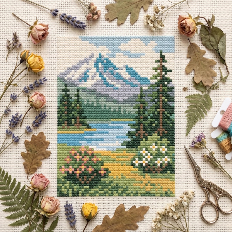 Puede incluir: Un bordado de punto de cruz que representa un paisaje de monta&ntilde;a con un lago, pinos y flores silvestres. La escena muestra monta&ntilde;as azules, un lago azul claro, &aacute;rboles verdes y un primer plano de hierba amarilla y verde con peque&ntilde;as flores blancas.