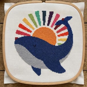 Op de afbeelding: Kruissteekpatroon met een blauwe walvis met een grijze buik en een regenboogzon. De walvis is geborduurd op witte stof, gehouden in een houten borduurring. De tekst onderaan luidt "CROSS STITCH PATTERN - INSTANT DOWNLOAD."