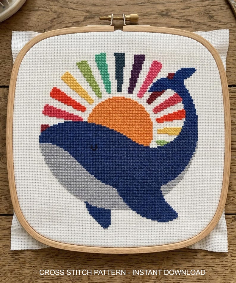 Op de afbeelding: Kruissteekpatroon met een blauwe walvis met een grijze buik en een regenboogzon. De walvis is geborduurd op witte stof, gehouden in een houten borduurring. De tekst onderaan luidt "CROSS STITCH PATTERN - INSTANT DOWNLOAD."