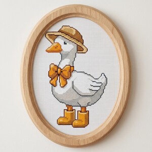 May include: Cross-stitch picture of a white goose wearing a brown hat, orange bow tie, and orange boots. The goose is framed in an oval wooden frame. The artwork is on a white background.