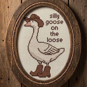 Op de afbeelding: Kruissteekkunstwerk van een gans met een cowboyhoed en laarzen, omlijst in een rustiek ovaal. De tekst luidt "silly goose on the loose" in bruine letters. Het frame is gemaakt van hout en touw.