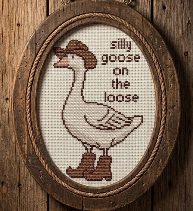 Op de afbeelding: Kruissteekkunstwerk van een gans met een cowboyhoed en laarzen, omlijst in een rustiek ovaal. De tekst luidt "silly goose on the loose" in bruine letters. Het frame is gemaakt van hout en touw.