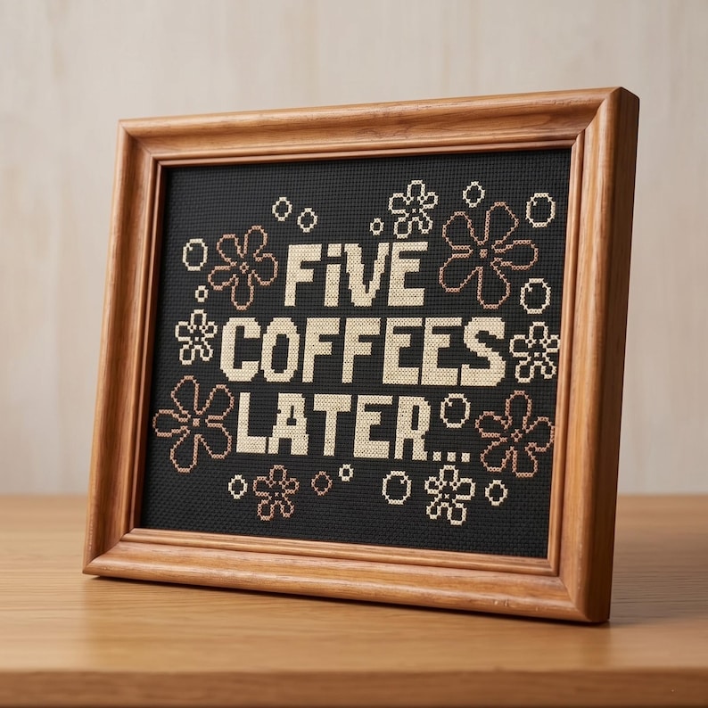 May include: A cross-stitch embroidery in a wooden frame featuring the text FIVE COFFEES LATER... in beige thread on a black fabric background, surrounded by small beige flower and circle motifs stitched in a grid pattern across the fabric.