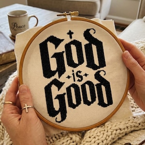 Peut inclure: Un tambour &agrave; broder circulaire en bois contenant un morceau de tissu Aida blanc cass&eacute;. Le tissu pr&eacute;sente une broderie au point de croix noire avec le texte "God is Good" dans une police gothique grasse, orn&eacute; de petites croix et d'&eacute;toiles.