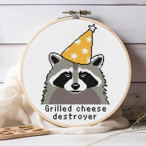 Op de afbeelding: Kruissteekborduurwerk van een wasbeer met een gele feesthoed met witte sterren. De wasbeer heeft grijze vacht, zwarte markeringen en de tekst "Grilled cheese destroyer" eronder. De borduursel zit in een houten borduurring.
