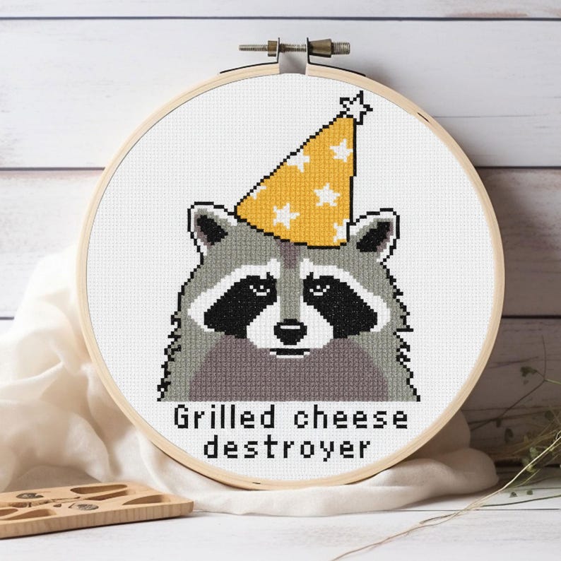 Op de afbeelding: Kruissteekborduurwerk van een wasbeer met een gele feesthoed met witte sterren. De wasbeer heeft grijze vacht, zwarte markeringen en de tekst "Grilled cheese destroyer" eronder. De borduursel zit in een houten borduurring.