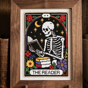 K&ouml;nnte beinhalten: Gerahmte Kreuzstichkunst mit einem Skelett, das ein Buch liest, mit der Aufschrift "THE READER". Das Kunstwerk hat einen schwarzen Hintergrund mit Sternen, einem Mond und bunten Blumen. Der Rahmen ist aus dunklem Holz.