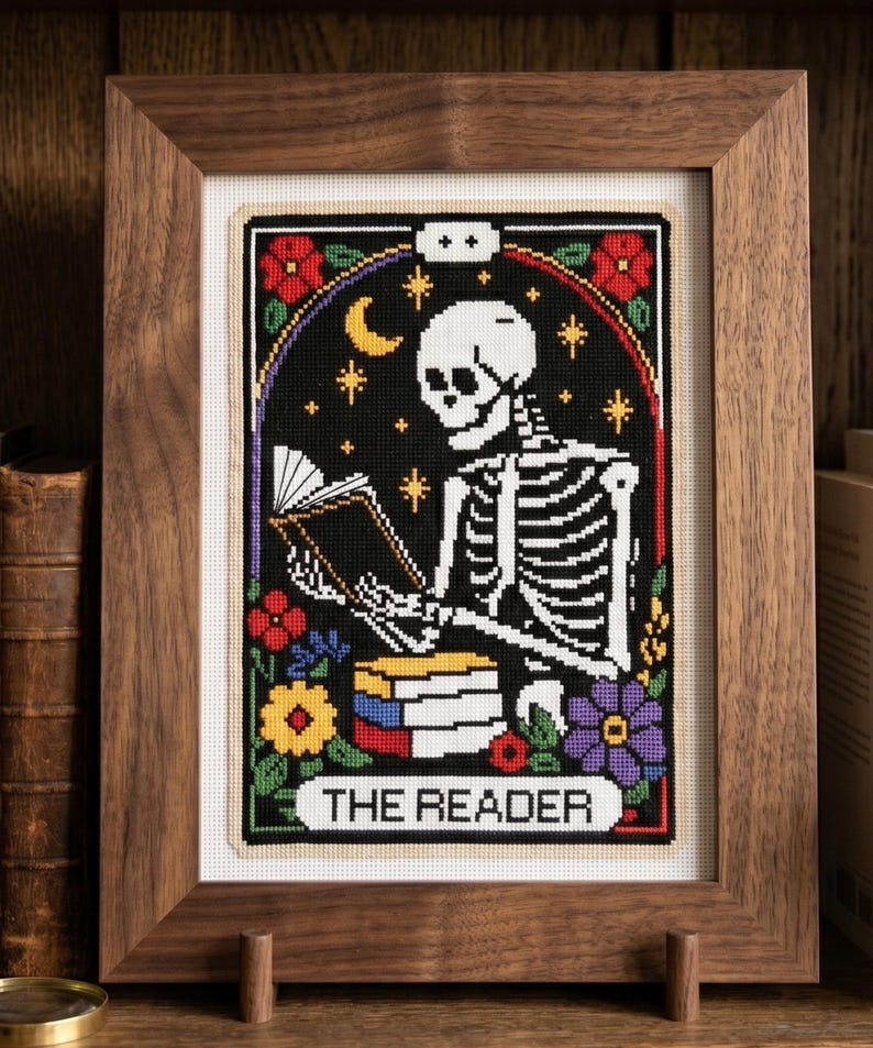 K&ouml;nnte beinhalten: Gerahmte Kreuzstichkunst mit einem Skelett, das ein Buch liest, mit der Aufschrift "THE READER". Das Kunstwerk hat einen schwarzen Hintergrund mit Sternen, einem Mond und bunten Blumen. Der Rahmen ist aus dunklem Holz.